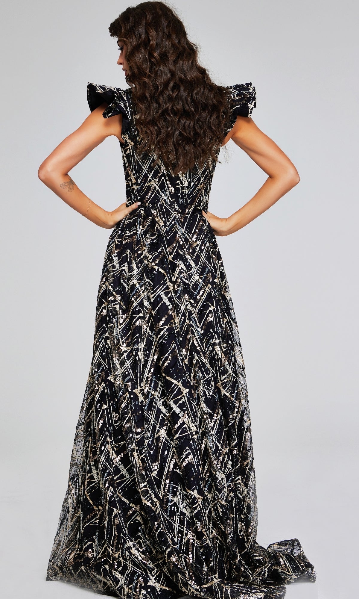 A woman stands with hands on hips, her back to the camera, modeling the Long Formal Dress: Jovani 39274โa striking black and gold evening gownโagainst a plain background.